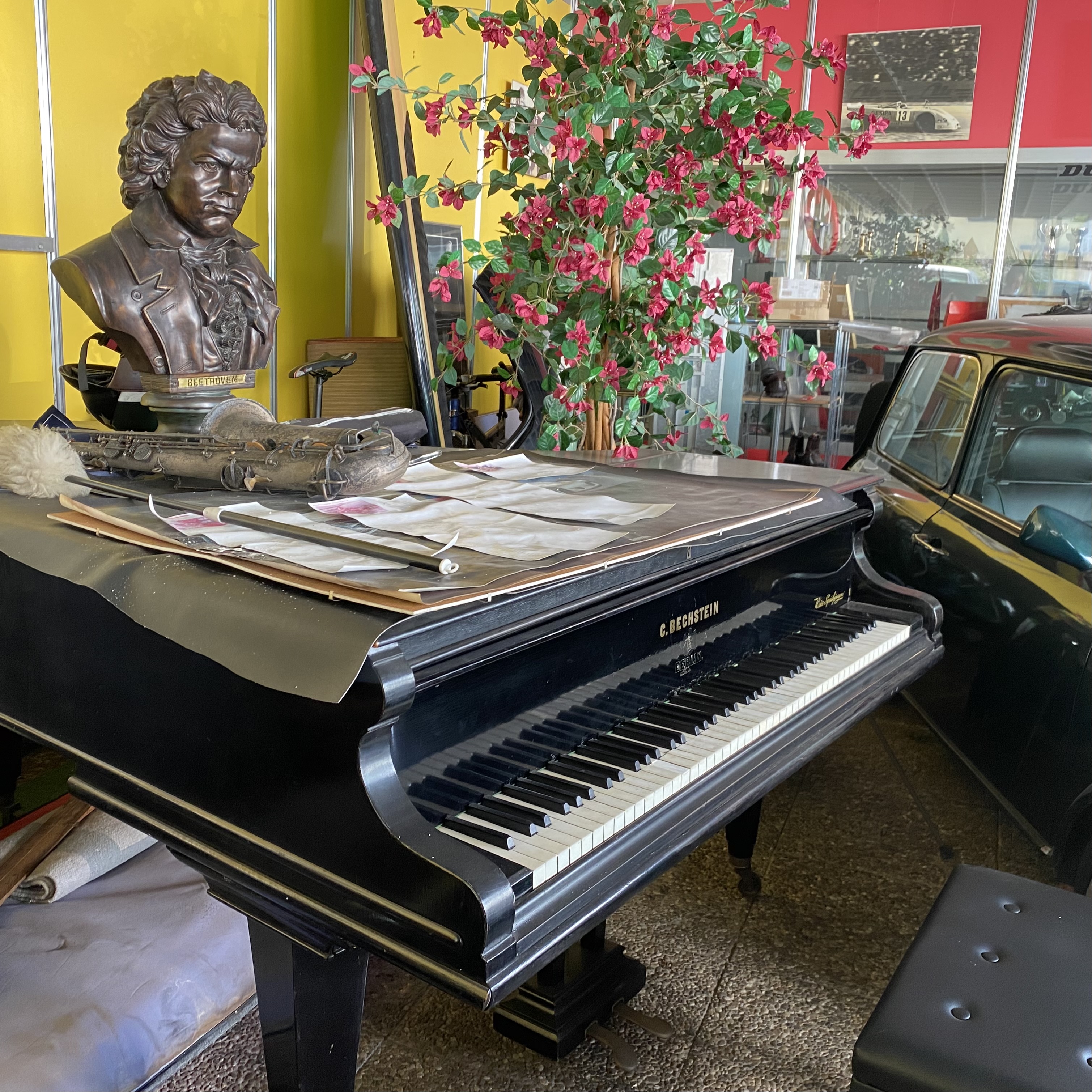 A black grand piano with sheet music and an antique saxophone on top, accompanied by a bust of Beethoven and a flowering plant in the background.