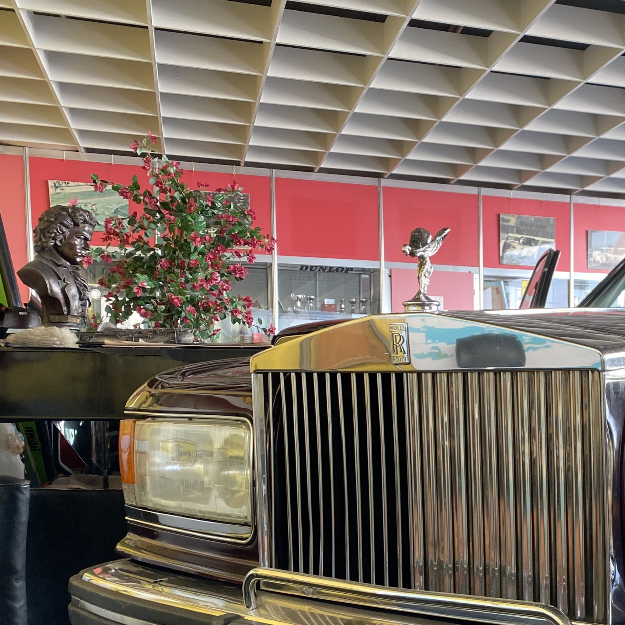 Close-up view of a Rolls-Royce car's front with a detailed grille and hood ornament. In the background, there is a bust of a composer and a floral arrangement.
