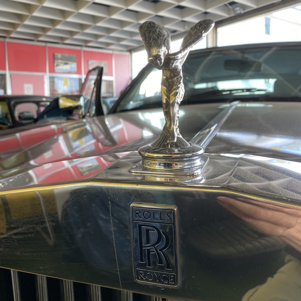 Close-up of a Rolls-Royce hood ornament, featuring the iconic Spirit of Ecstasy figurine, on the front of a luxury vehicle in a showroom.