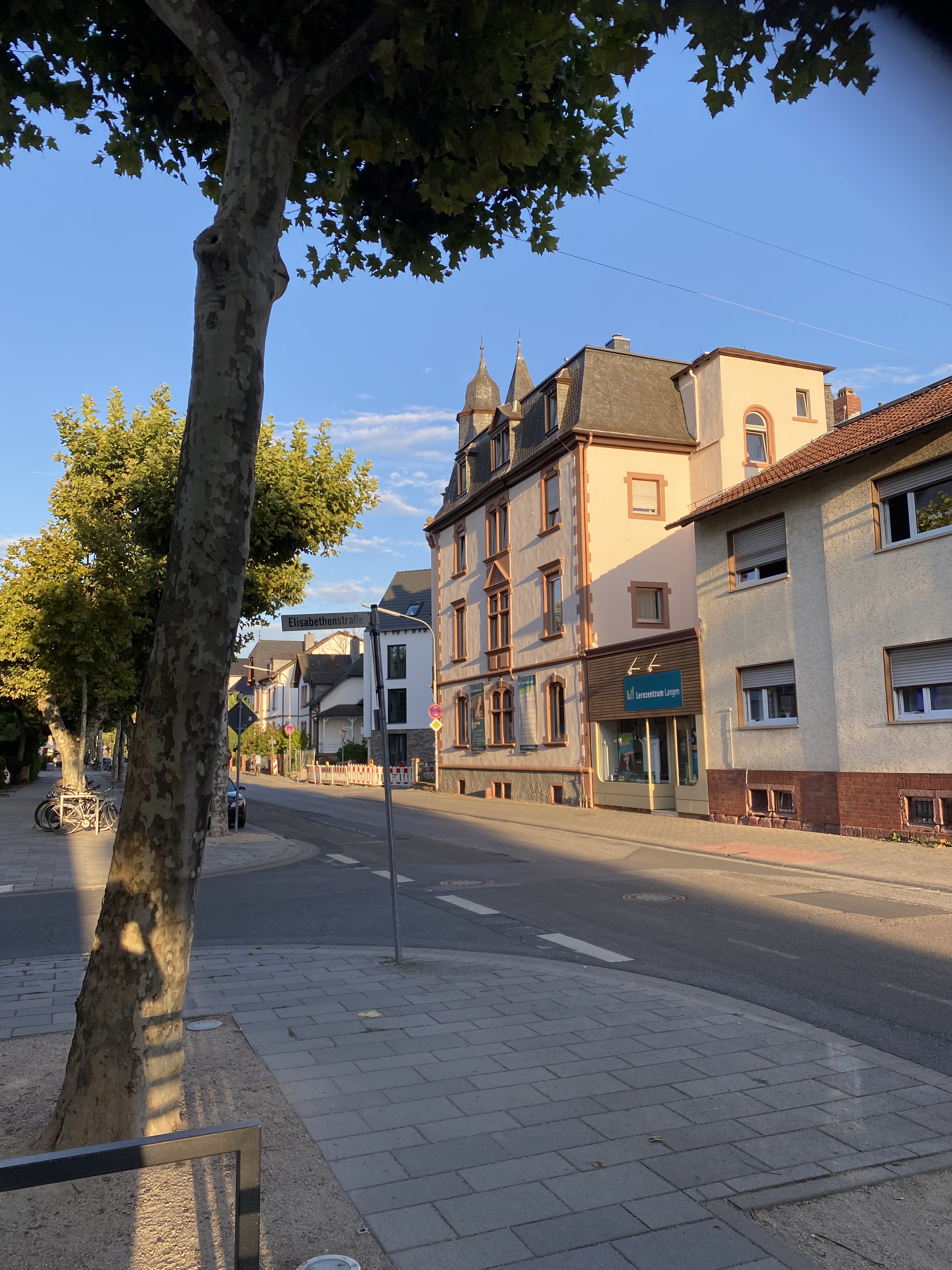 A street view featuring a tree and several buildings, including a pink multi-story building with unique architectural features and a neighboring property. The sky is clear with a hint of clouds.