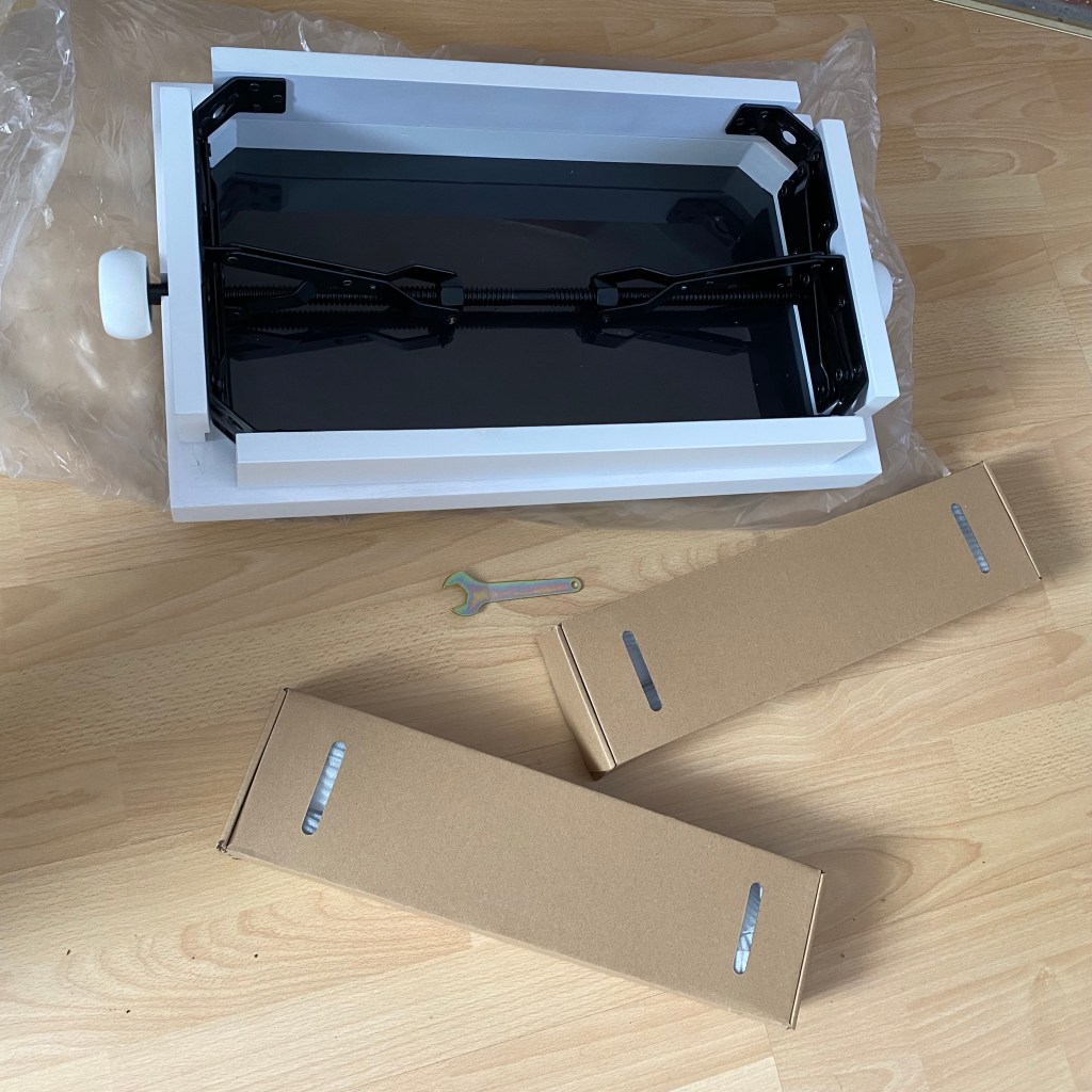 An unassembled product with a black top part, white sides, two brown cardboard boxes, a small tool, and plastic wrapping on a wooden surface.