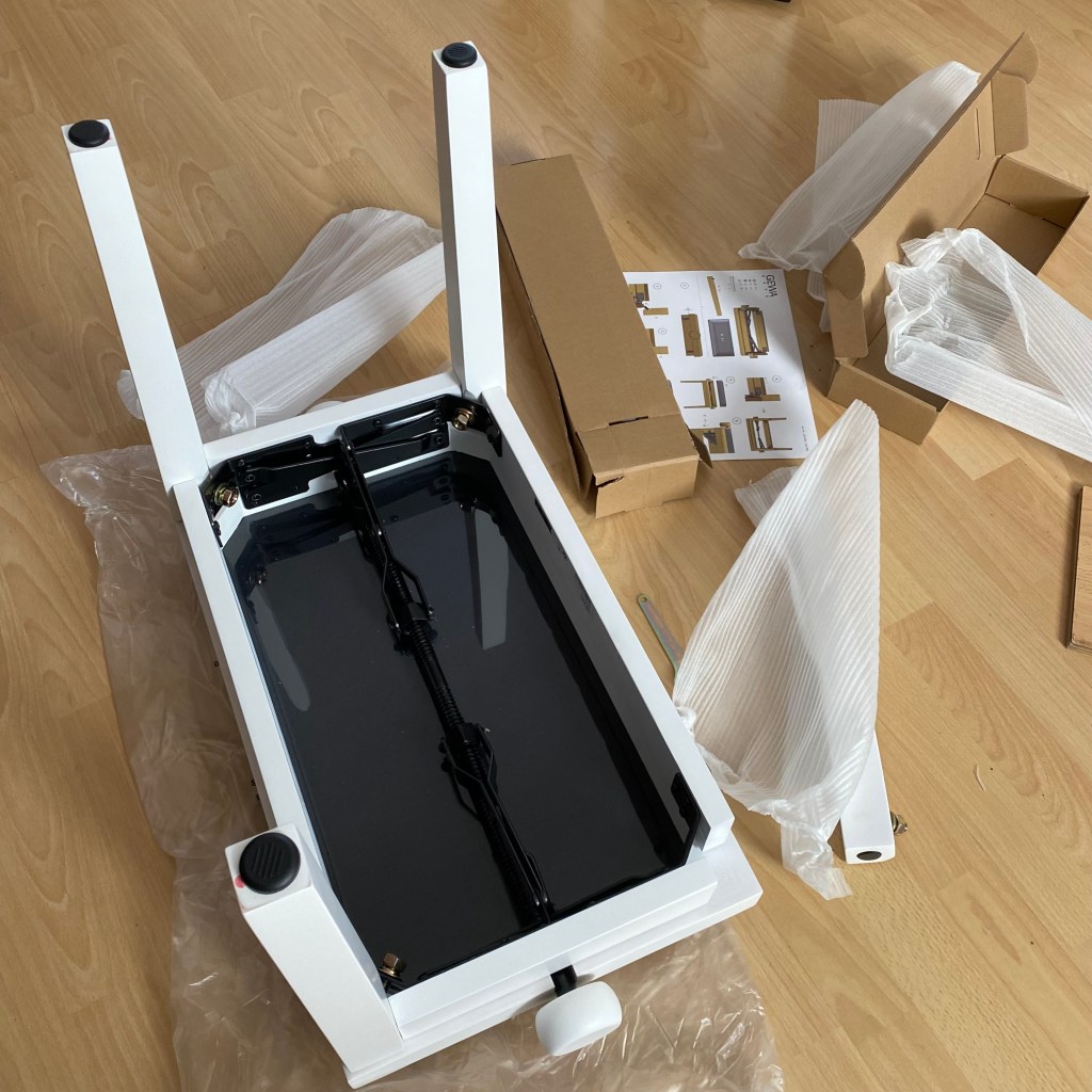 An unassembled furniture piece with a white frame and a black base, surrounded by packaging materials and assembly instructions on a wooden floor.