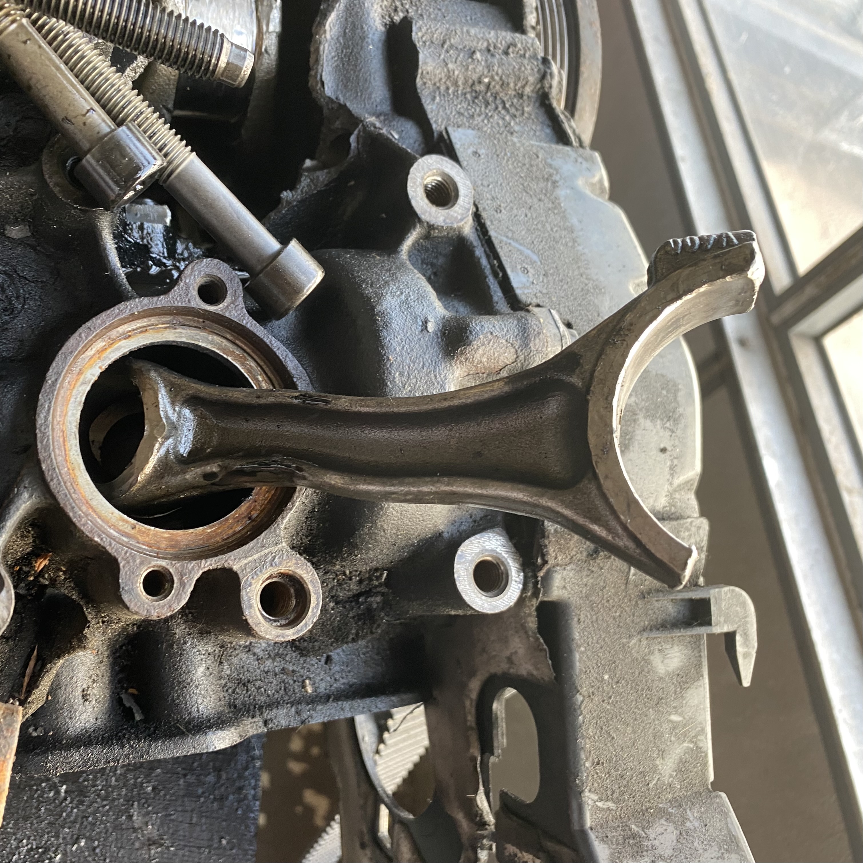 Close-up of a metallic engine component, specifically a connecting rod mounted on a dark engine block with visible bolts and oil residue.