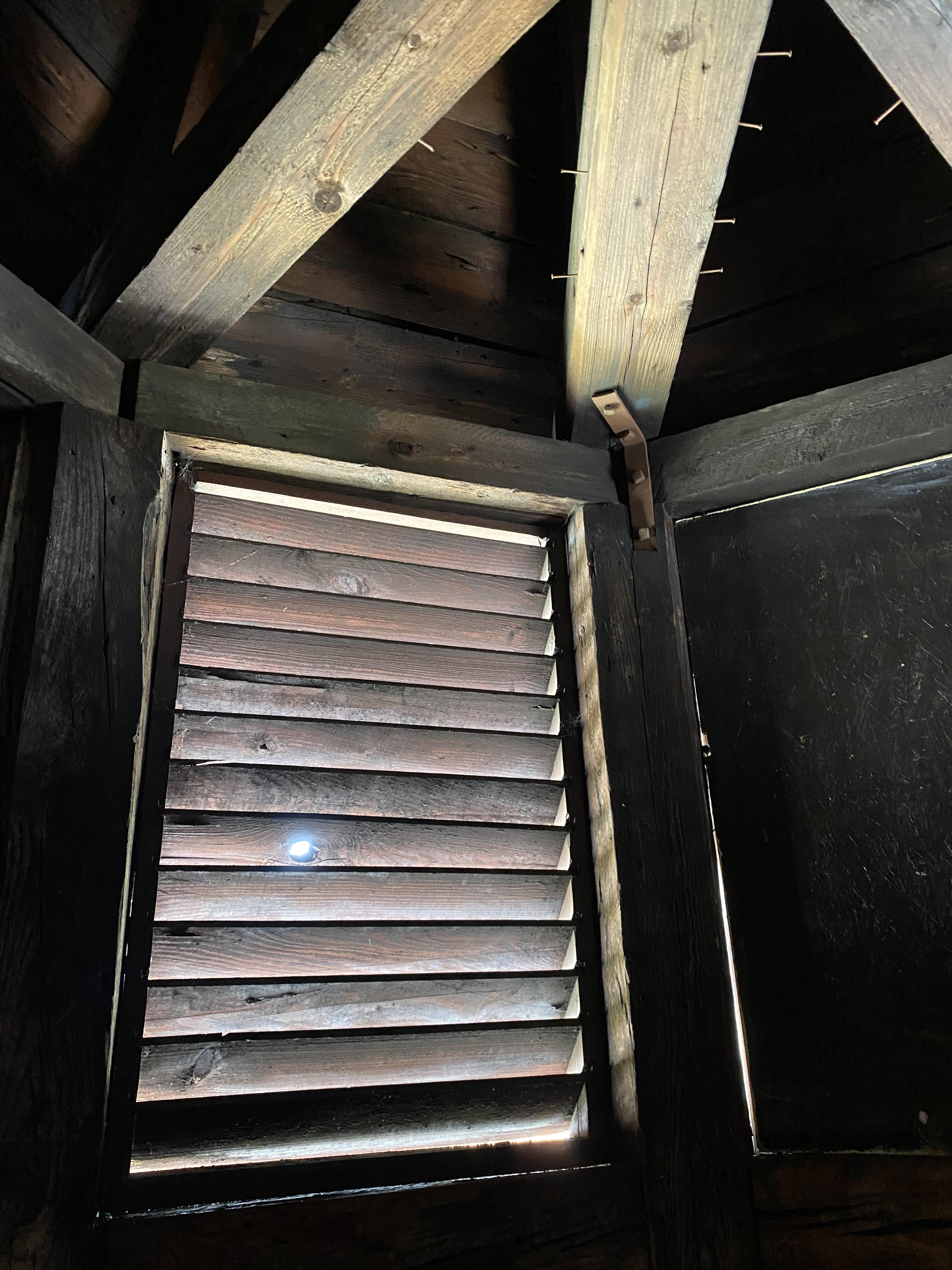 View of a wooden interior with beams and a partially closed shutter, allowing light to filter through a small opening.