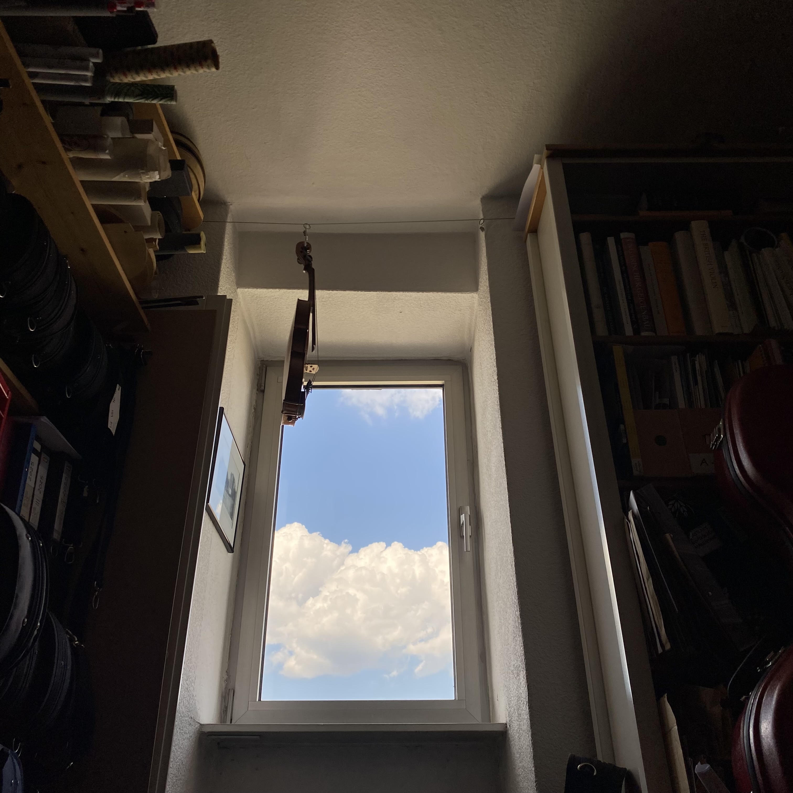 A view from inside a room looking out of a window, revealing a bright blue sky with clouds. A violin is positioned above the window, and bookshelves are visible in the background.