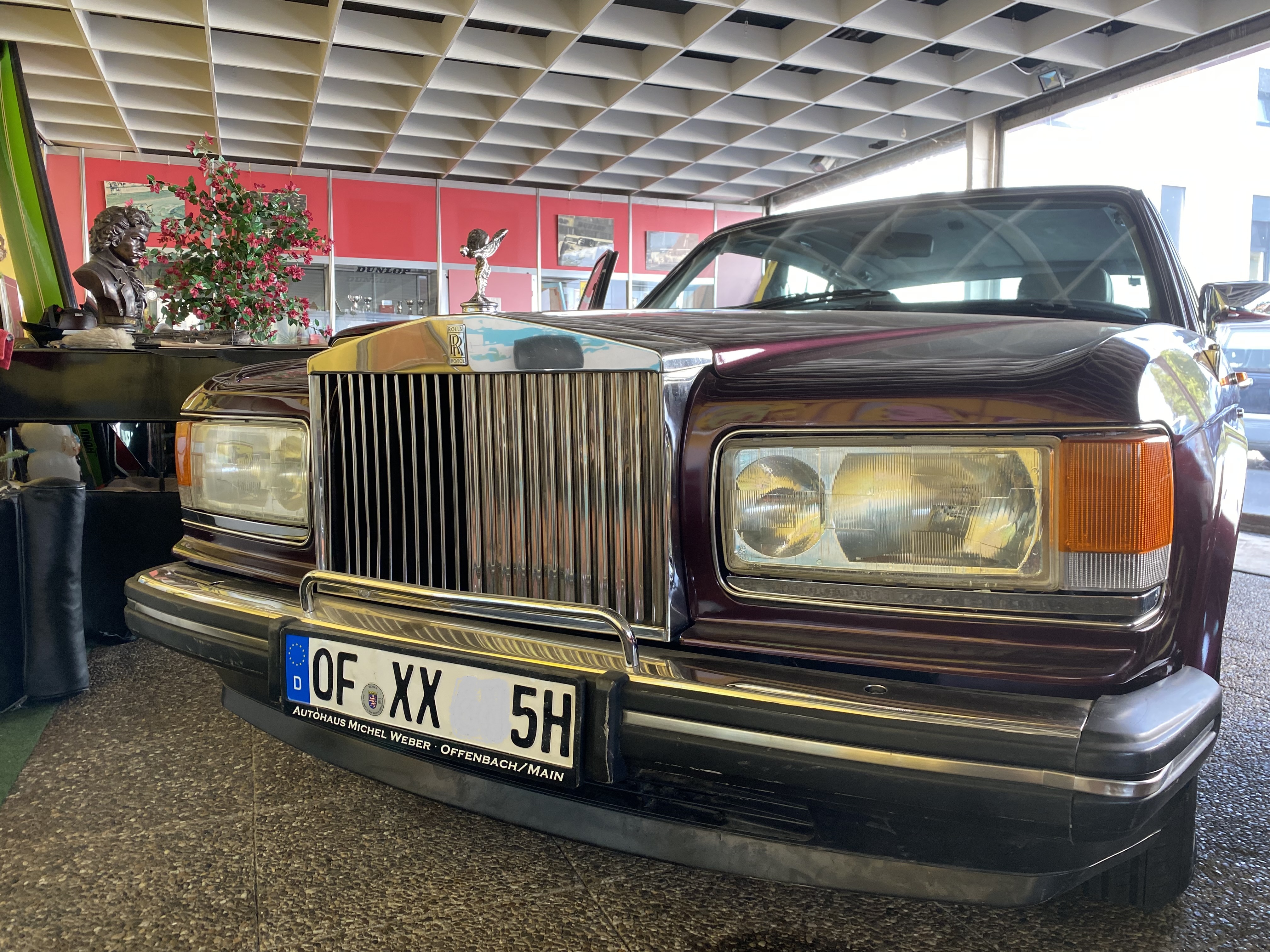 Front view of a vintage Rolls-Royce car with a shiny grille and distinctive hood ornament, parked in an indoor setting with decorative items in the background.