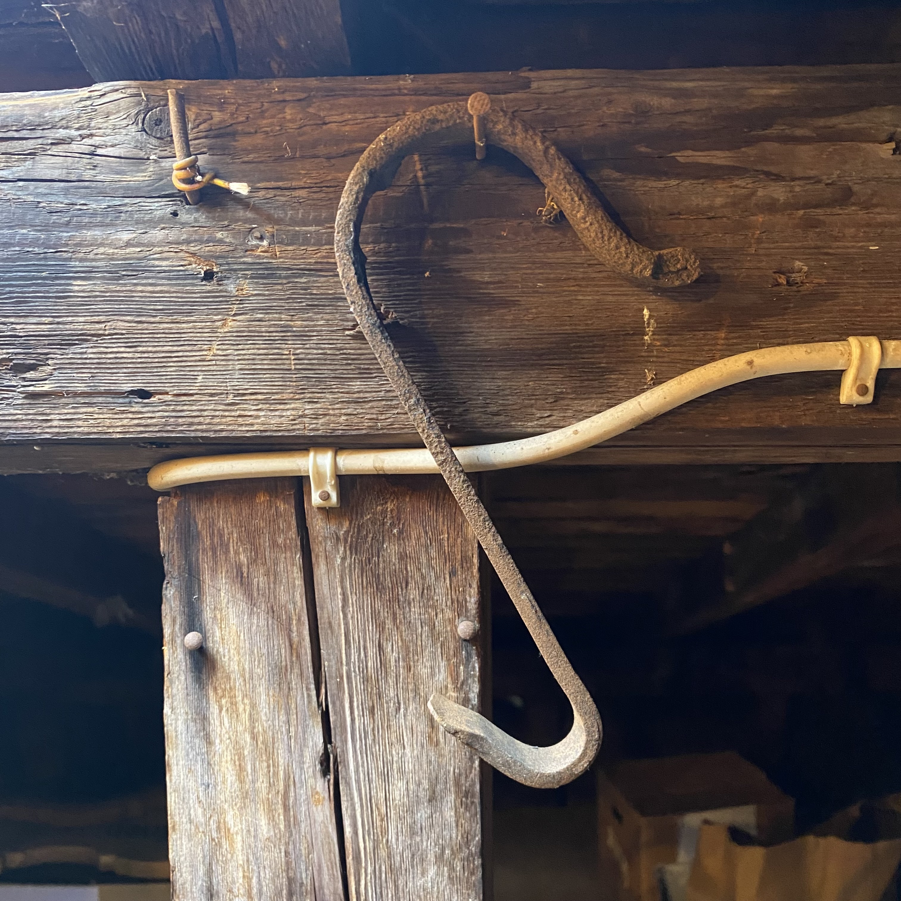 An old, rusted metal hook hanging on a wooden beam, with a white plastic pipe mounted below it. The background features dark wood and boxes.