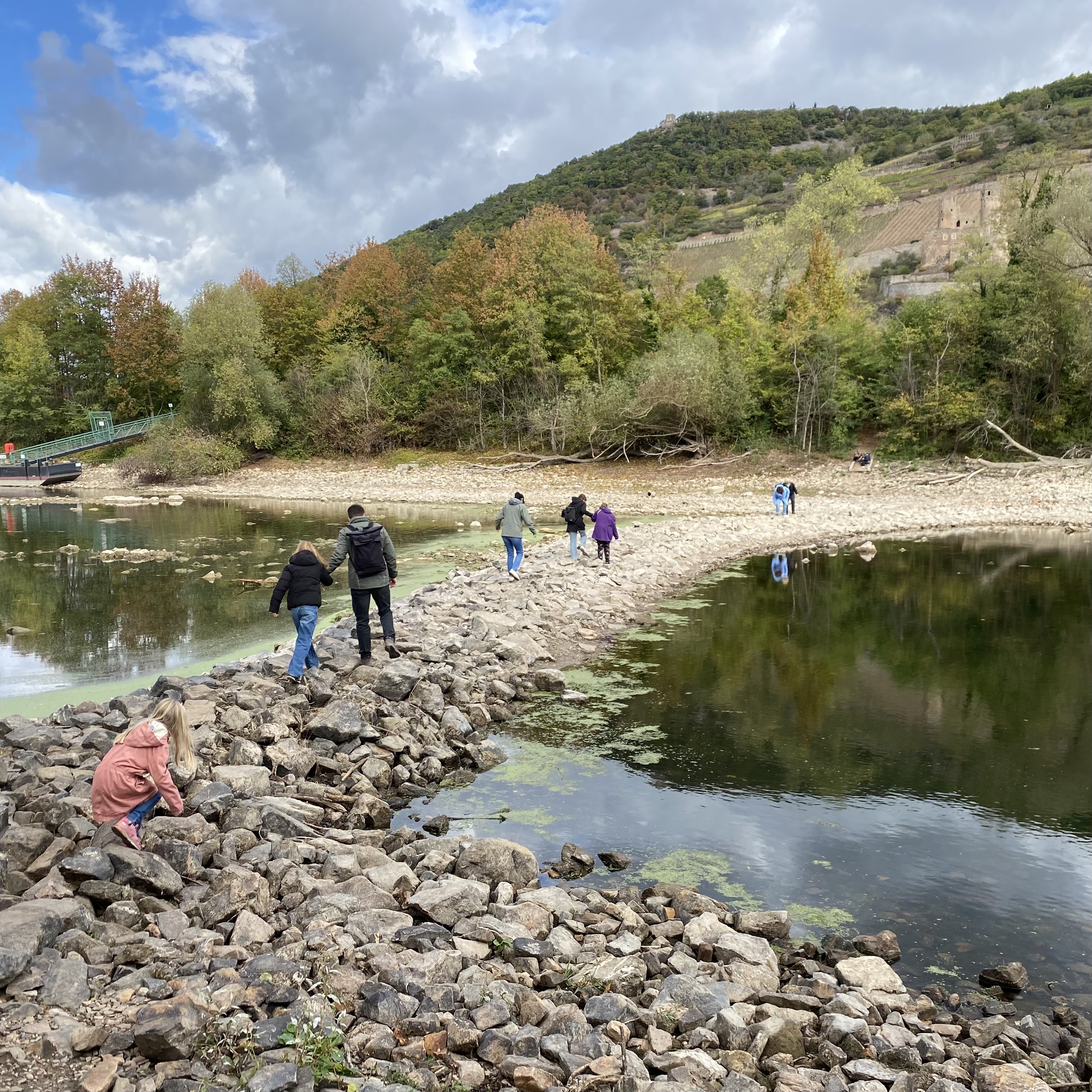 People walking on a rocky path along a riverbank, surrounded by trees with autumn foliage and a cloudy sky.