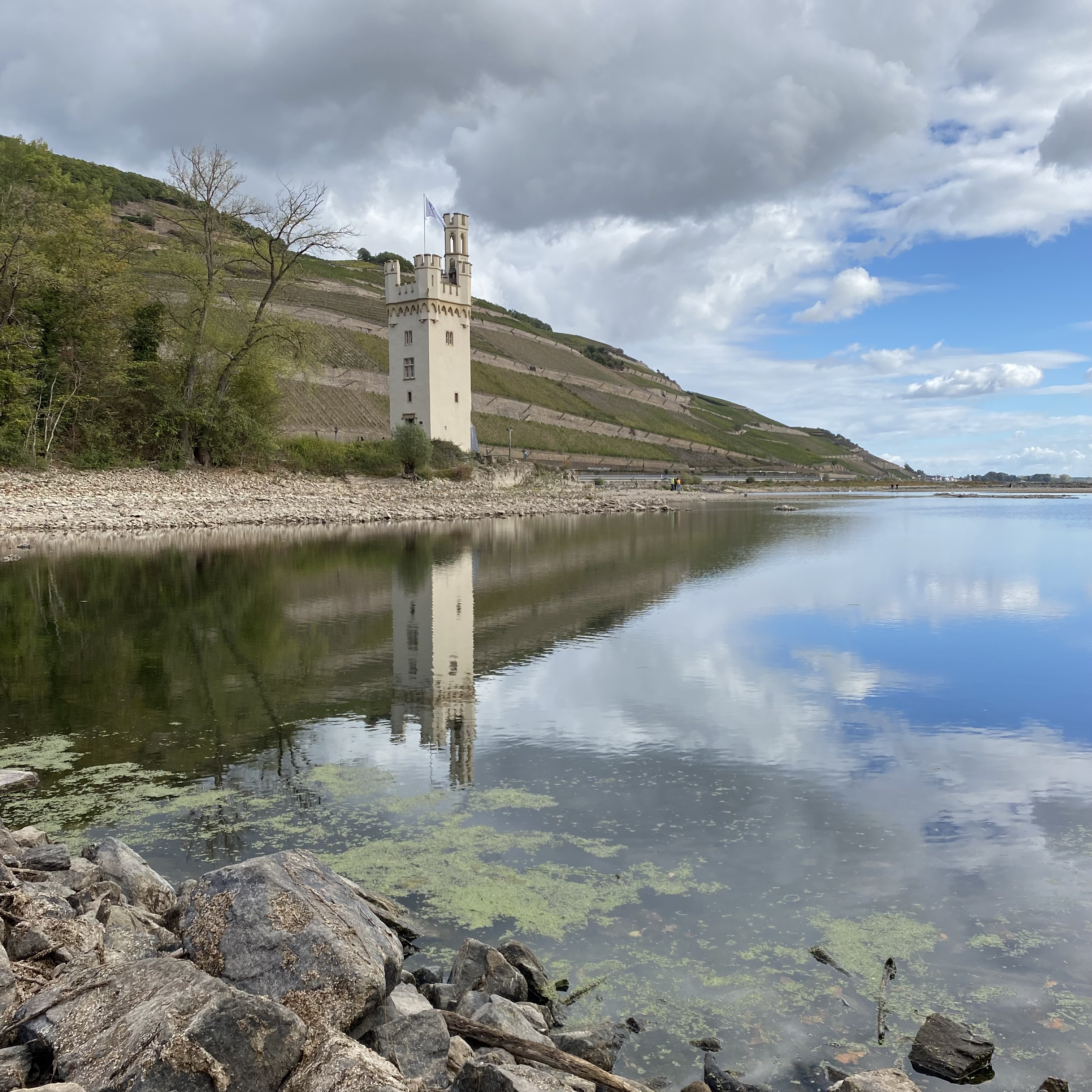 A scenic view of a white castle tower reflected in a calm river, surrounded by greenery and hills under a partly cloudy sky.