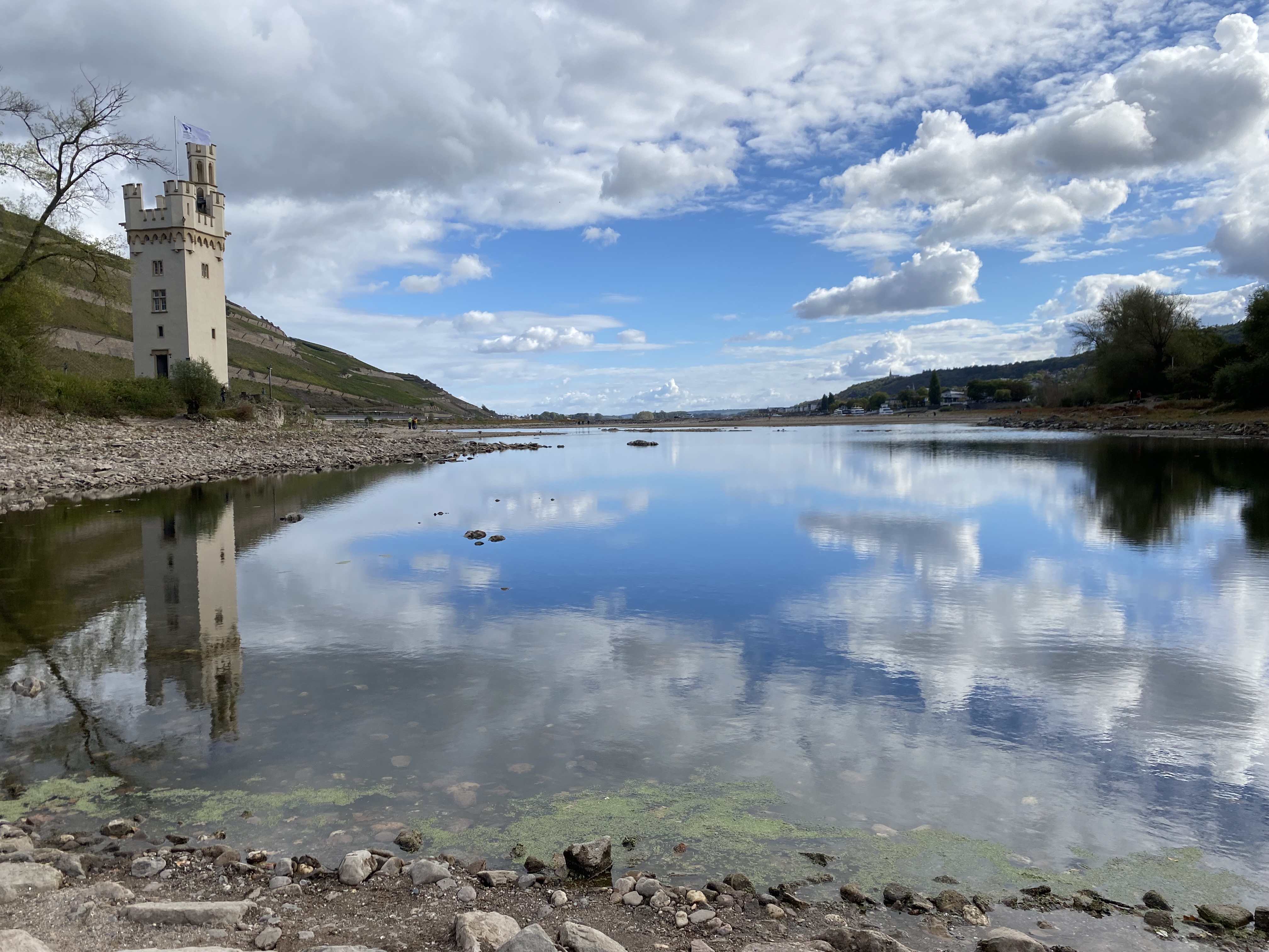 A tall, white tower reflecting in a calm body of water, surrounded by green hills and clouds in the blue sky.