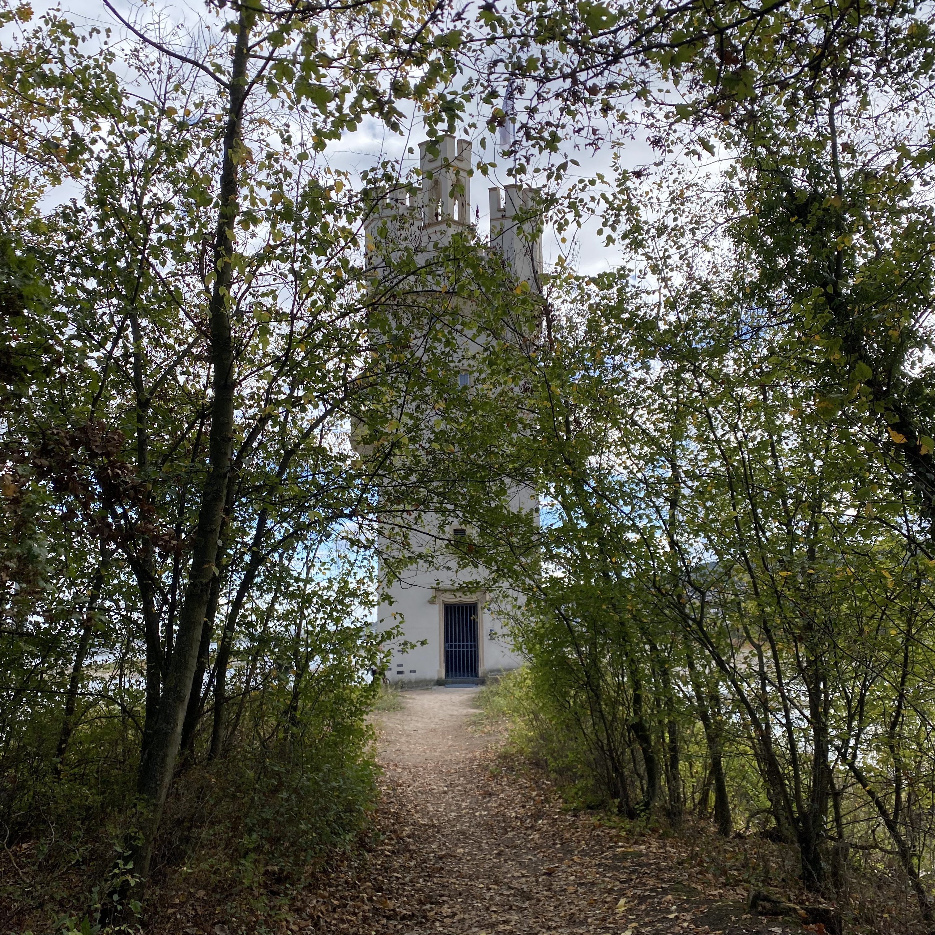 A pathway surrounded by trees leading to a white tower in the distance under a cloudy sky.