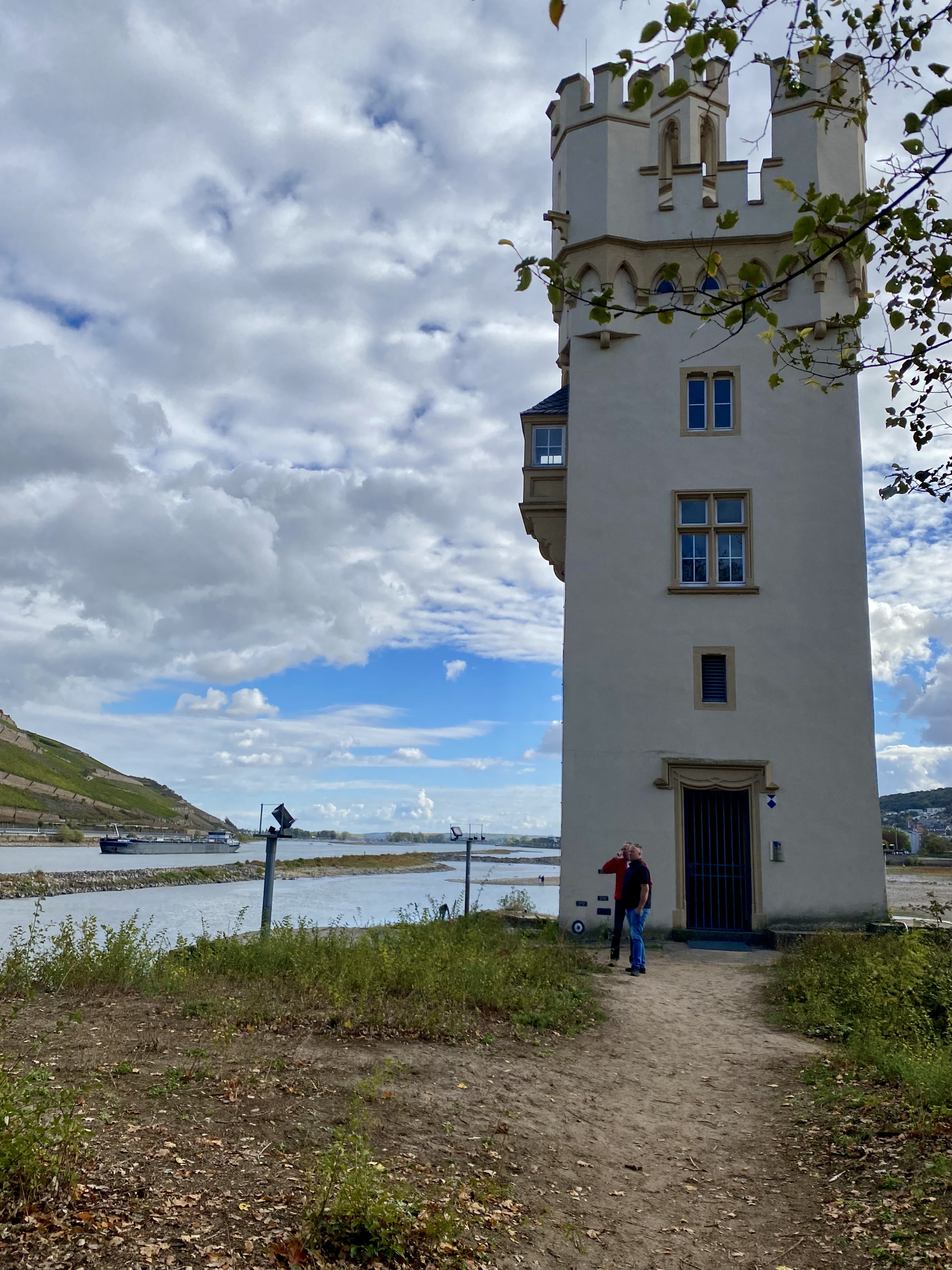 A narrow white tower with a castle-like design stands near a river, surrounded by greenery and under a cloudy sky.