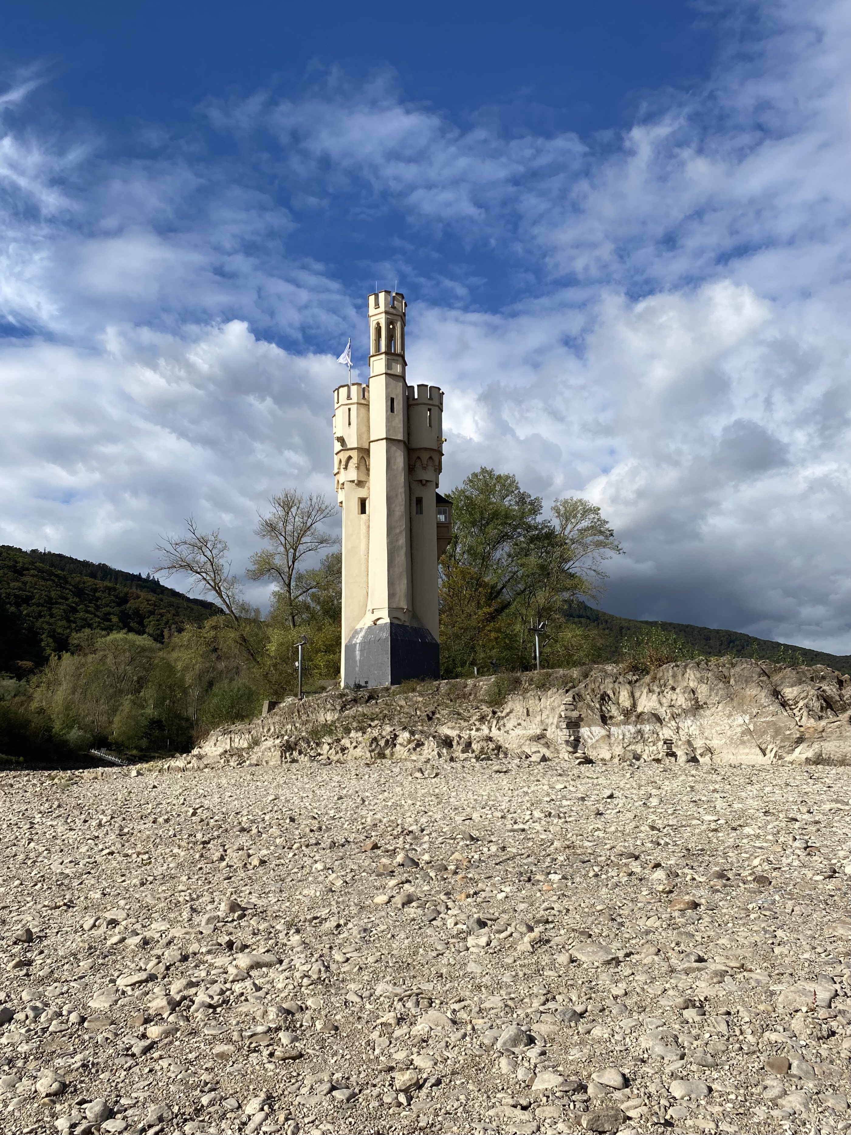 A tall, beige stone tower with a rounded top stands on a rocky shoreline under a blue sky with scattered clouds.