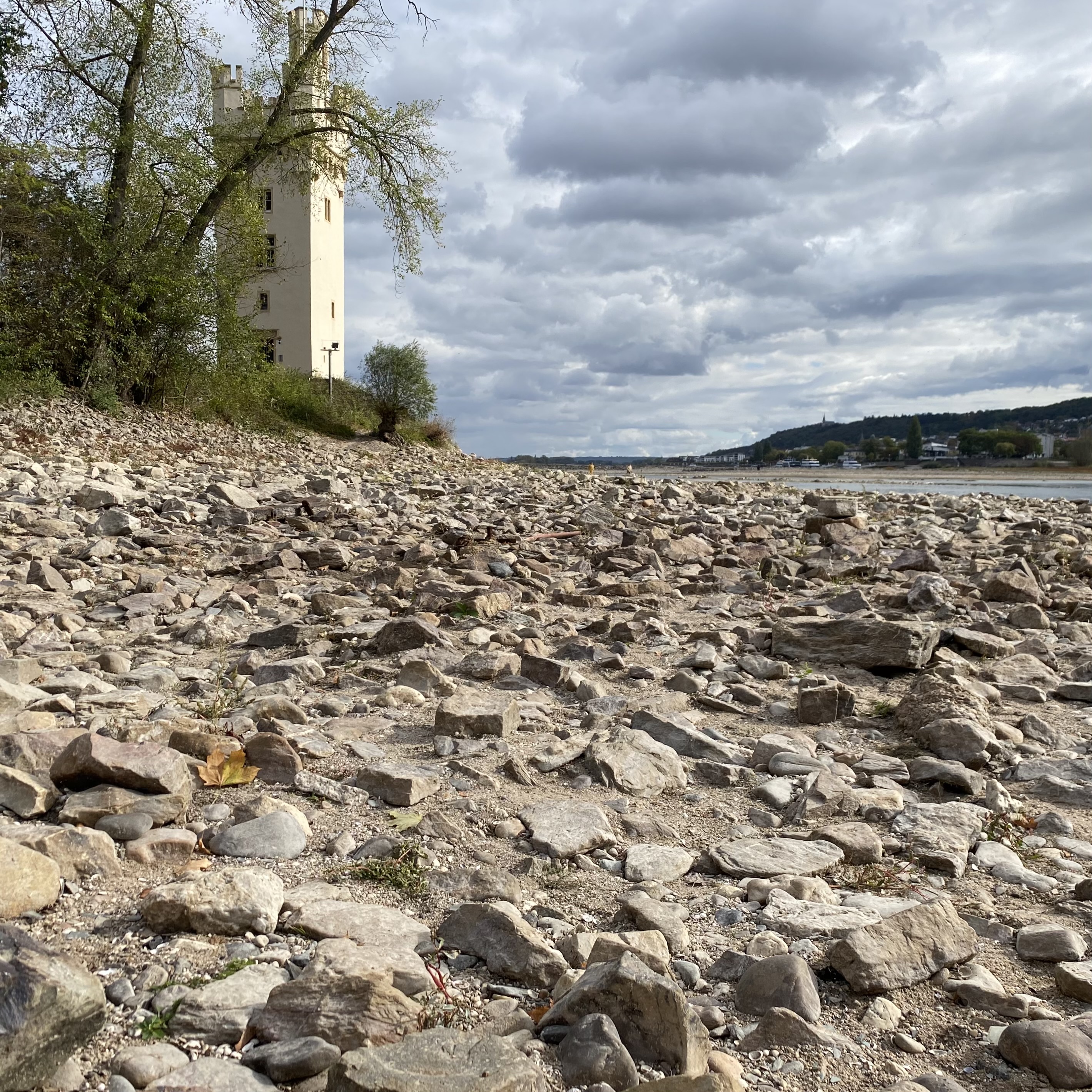 A rocky riverbank with a white tower in the background under a cloudy sky.