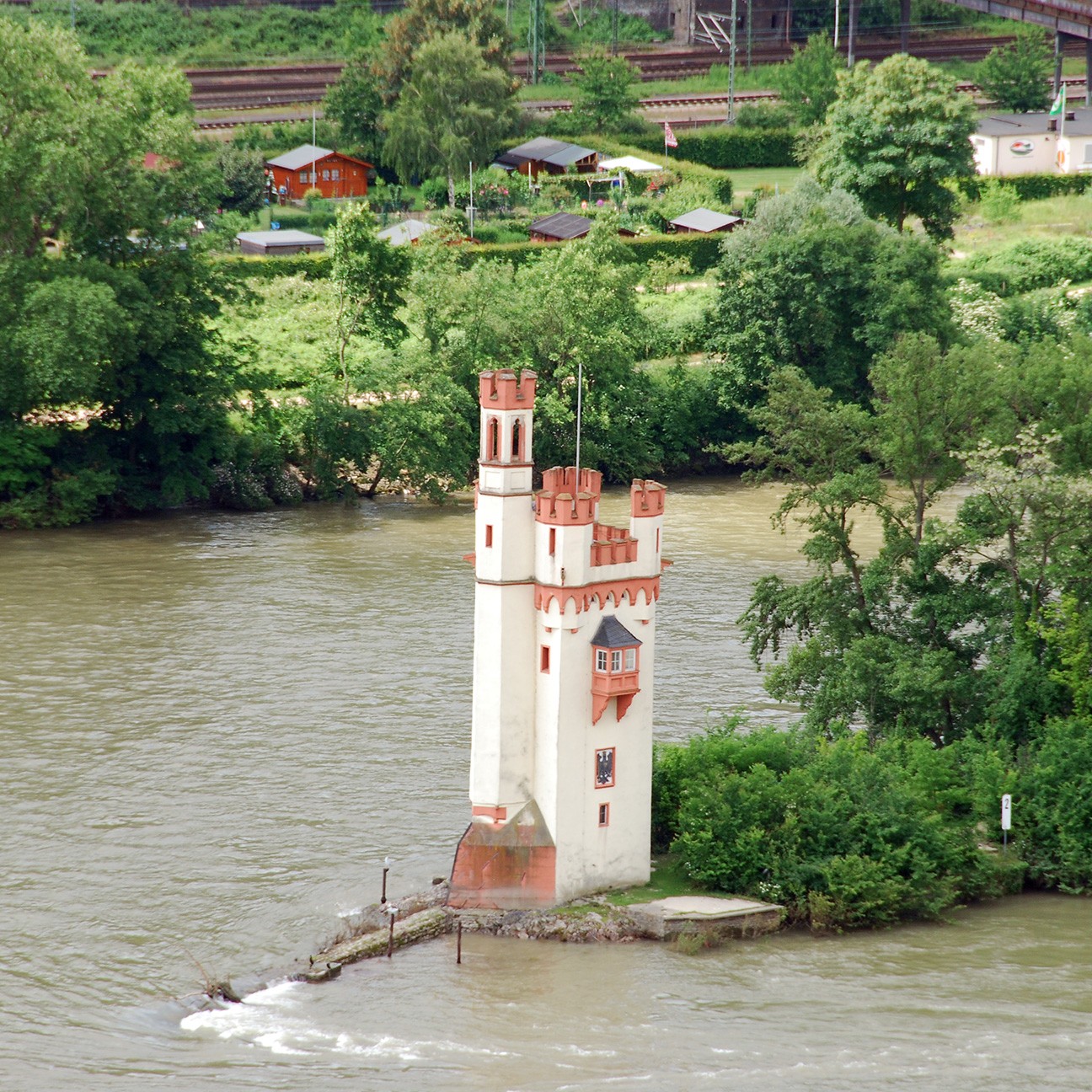 View of a decorative tower situated on a small island in a river, surrounded by greenery and trees.